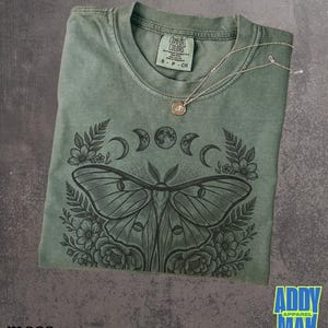 May include: Moss green Comfort Colors t-shirt with a moth and moon phases graphic. A silver necklace with a small pendant rests on the shirt. The shirt has a crew neck and short sleeves. The brand name "Addy Mak Apparel" is visible.