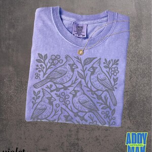 May include: Violet-colored t-shirt with a crew neck and a gray cardinal bird design. The shirt has a Comfort Colors tag and a gold pendant necklace. The text "Addy Mak" is in a blue and yellow box.