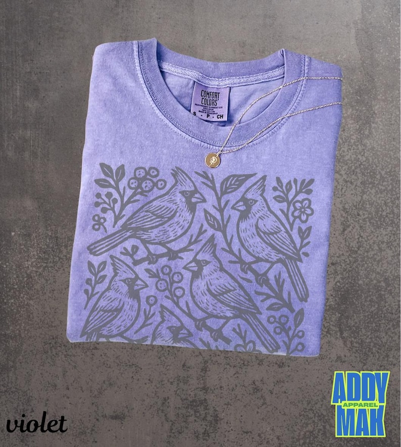 May include: Violet-colored t-shirt with a crew neck and a gray cardinal bird design. The shirt has a Comfort Colors tag and a gold pendant necklace. The text "Addy Mak" is in a blue and yellow box.
