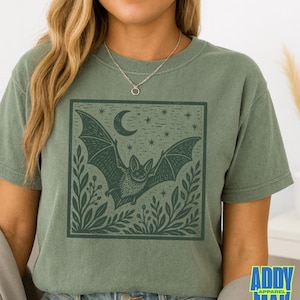 May include: A sage green short-sleeve t-shirt featuring a dark green square graphic print of a bat in flight, surrounded by foliage, a crescent moon, and stars. The shirt has a crew neckline and a relaxed fit, showcasing a detailed illustration.