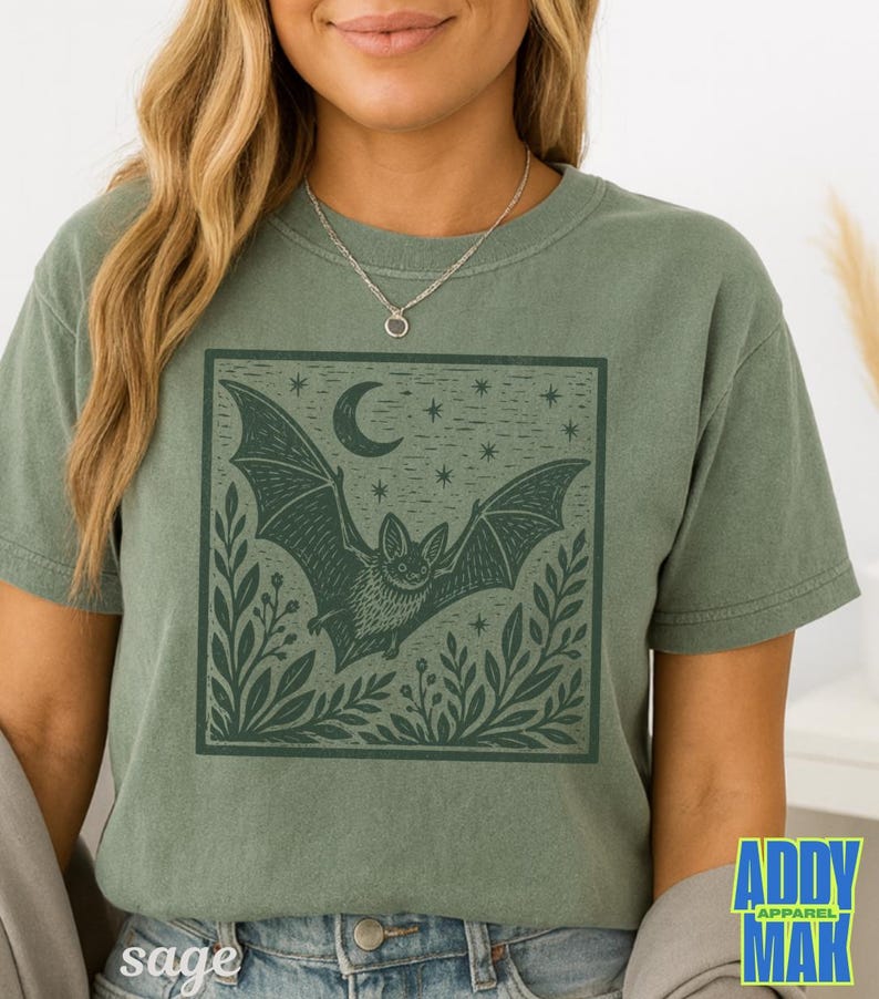 May include: A sage green short-sleeve t-shirt featuring a dark green square graphic print of a bat in flight, surrounded by foliage, a crescent moon, and stars. The shirt has a crew neckline and a relaxed fit, showcasing a detailed illustration.