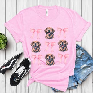 Puede incluir: Una camiseta rosa claro con un estampado de caras de golden retriever y lazos rosas. La camiseta se combina con zapatillas deportivas blancas y negras, pantalones cortos vaqueros y una peque&ntilde;a planta en maceta sobre una superficie de madera blanca.