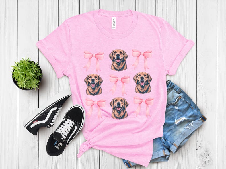 Puede incluir: Una camiseta rosa claro con un estampado de caras de golden retriever y lazos rosas. La camiseta se combina con zapatillas deportivas blancas y negras, pantalones cortos vaqueros y una peque&ntilde;a planta en maceta sobre una superficie de madera blanca.