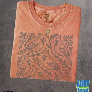May include: Coral-colored t-shirt with a gray cardinal bird design. The shirt features a crew neck and a tag that reads "Comfort Colors." A gold necklace with a pendant rests on the shirt. The brand "Addy Max Apparel" is visible.