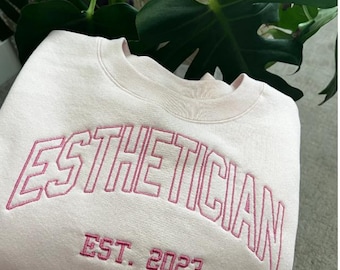 May include: A light pink sweatshirt with the word "ESTHETICIAN" embroidered in pink, along with "EST. 2023". The sweatshirt is made of a soft material and is folded. Green leaves are in the background.