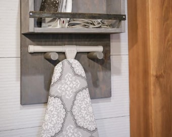 May include: A gray wooden wall-mounted ironing board holder with a shelf above. The shelf has a gray iron and a cord on it. The ironing board cover has a white and gray floral pattern.