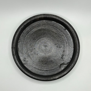May include: A black ceramic plate with a slightly raised rim. The plate has a smooth, slightly textured surface.