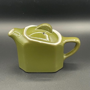 May include: Olive green ceramic teapot with a white rim around the lid. The teapot has a handle and a spout. The lid has a small handle. The teapot is a hexagonal shape.