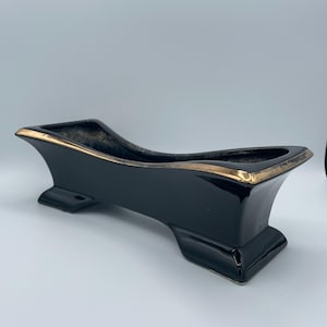 May include: A black ceramic planter with a gold trim. The planter has a rectangular shape with a curved top and a pedestal base. The interior is a dark, matte color.