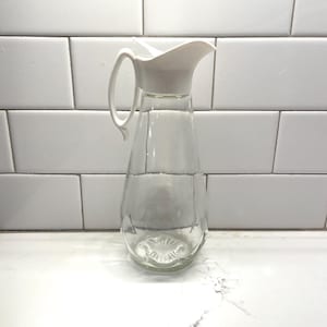 May include: A clear glass water pitcher with a white plastic spout and handle. The pitcher has a rounded base and a slender body. The handle is curved and attached to the side of the pitcher. The pitcher is sitting on a white surface.