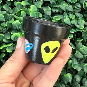 May include: A small, black painted ceramic pot with a heart and teacup design on one side and a yellow alien face on the other. The pot is held against a backdrop of green foliage.