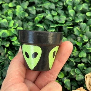 May include: A small black terracotta planter with green alien faces painted on the front. The planter is being held in a hand.