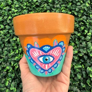 Puede incluir: Una maceta de terracota pintada con un ojo azul dentro de un corazón rosa. El corazón está rodeado por un borde azul y turquesa. La maceta está sentada en una mano.