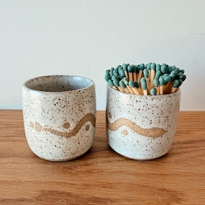 May include: Two speckled ceramic match holders with a wavy gold design. One holder is empty, and the other is filled with green-tipped matches. The holders are on a wooden surface.