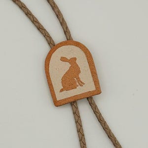 May include: A bolo tie with a rectangular pendant featuring a rabbit silhouette. The pendant is a light brown color with a cream-colored background. The braided cord is a light brown color.