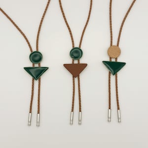 May include: Three bolo ties with geometric designs. Each tie features a dark green or brown triangle or circle pendant, paired with a matching circle. The pendants are attached to a brown cord with silver-colored tips. The background is white.