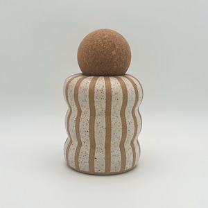 Puede incluir: Un juego de salero y pimentero de cerámica con un diseño de rayas blancas y marrones. La parte superior del salero es un pomo redondo de corcho.