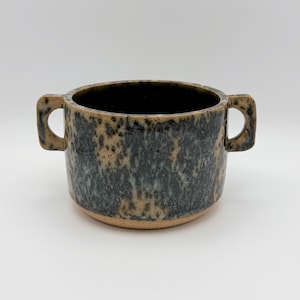 May include: A ceramic soup bowl with two side handles, featuring a speckled glaze in shades of dark charcoal grey and tan. The bowl has a wide, cylindrical body with a smooth rim and a visible unglazed tan base at the bottom of the vessel.