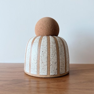 May include: A ceramic jar with a dome-shaped lid featuring a textured brown cork-like finish. The base is a rounded cylinder with alternating vertical stripes of speckled off-white glaze and smooth, unglazed light brown clay.