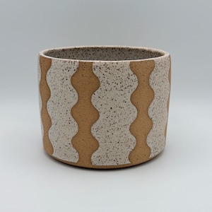 Peut inclure: Un pot de fleurs cylindrique en céramique avec une finition blanc cassé mouchetée contrastée par des bandes verticales ondulées en argile beige. Le récipient présente un profil droit et lisse avec une large ouverture et une base plate.