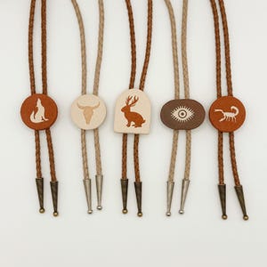 May include: A collection of bolo ties with various designs. The ties feature leather cords and decorative slides with images of a wolf, bull skull, deer, eye, and scorpion. The slides are in shades of brown and cream.
