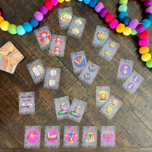 May include: A set of colorful cards with illustrations of religious symbols and text. The cards are laid out on a wooden table with a rainbow garland in the background.