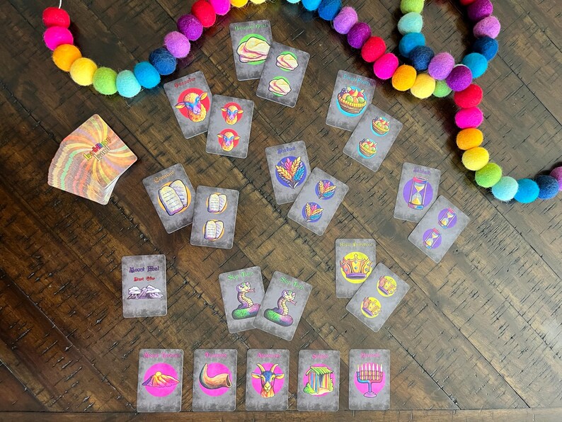 May include: A set of colorful cards with illustrations of religious symbols and text. The cards are laid out on a wooden table with a rainbow garland in the background.