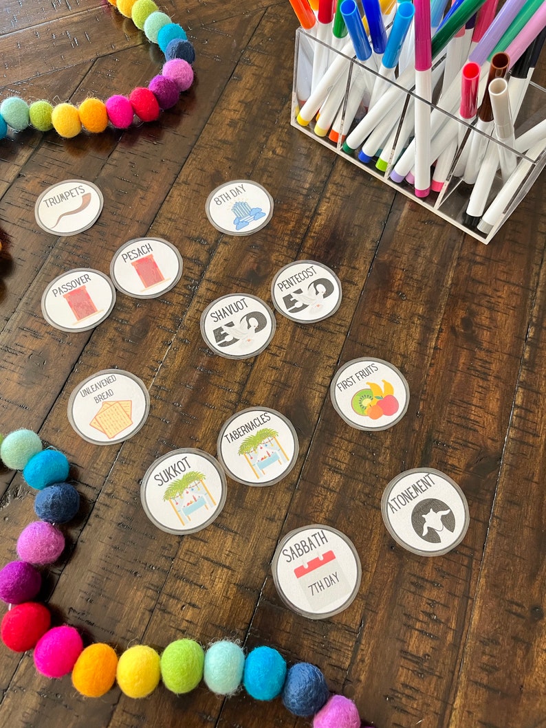 May include: A set of colorful, round, illustrated stickers with Jewish holidays names and symbols. The stickers include Passover, Pesach, Unleavened Bread, Shavuot, Pentecost, Sukkot, Tabernacles, First Fruits, Sabbath, 7th Day, Atonement, and Trumpets. The stickers are on a wooden table with a string of colorful pom-poms.