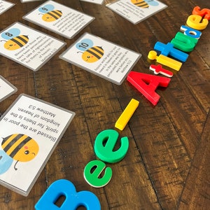 Puede incluir: Un conjunto de diez tarjetas con una ilustraci&oacute;n de una abeja y un vers&iacute;culo b&iacute;blico sobre las Bienaventuranzas. Las tarjetas est&aacute;n sobre una mesa de madera con la palabra "Bee-Attitudes" escrita en letras magn&eacute;ticas de colores.