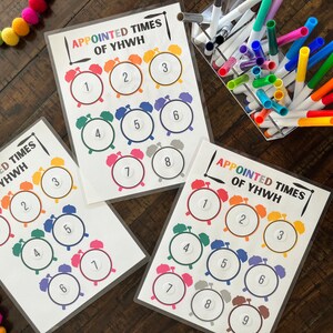 May include: Three printable sheets with the text "Appointed Times of YHWH" and colorful alarm clock images with numbers 1 through 9. The sheets are on a wooden surface with a container of colorful dry-erase markers.