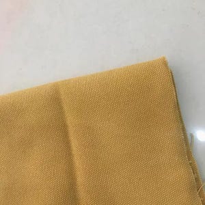 Op de afbeelding: Close-up van een gedeelte van mosterdgeel canvasweefsel. De stof heeft een strakke weefsel en een licht gestructureerd oppervlak. De randen van de stof zijn zichtbaar en tonen de rauwe, onafgewerkte rand.