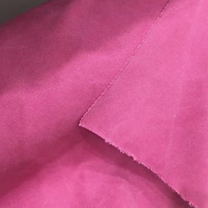 Op de afbeelding: Close-up van een felroze stof met een geweven textuur. De stof is gevouwen, waardoor een rafelige rand en een naad zichtbaar zijn. De heldere kleur en textuur suggereren gebruik voor kleding of interieurdecoratie.