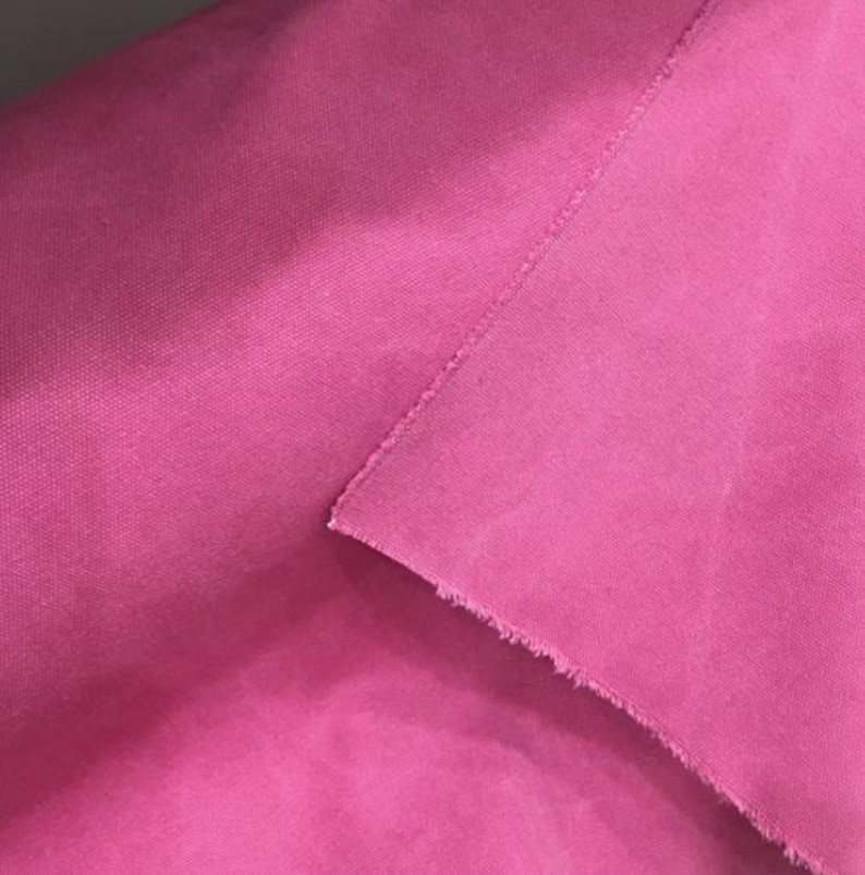 Op de afbeelding: Close-up van een felroze stof met een geweven textuur. De stof is gevouwen, waardoor een rafelige rand en een naad zichtbaar zijn. De heldere kleur en textuur suggereren gebruik voor kleding of interieurdecoratie.