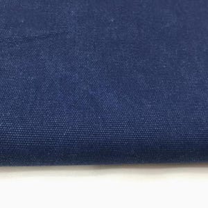 Op de afbeelding: Close-up van een gedeelte van marineblauwe canvasstof. Het strak geweven materiaal heeft een getextureerd uiterlijk, met zichtbare kleine, donkerblauwe vezels. De stof is omgevouwen, waardoor een witte rand aan de onderkant zichtbaar is.