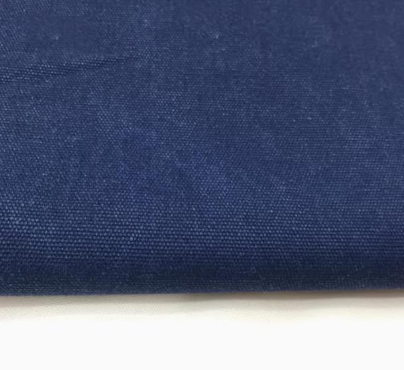 Op de afbeelding: Close-up van een gedeelte van marineblauwe canvasstof. Het strak geweven materiaal heeft een getextureerd uiterlijk, met zichtbare kleine, donkerblauwe vezels. De stof is omgevouwen, waardoor een witte rand aan de onderkant zichtbaar is.