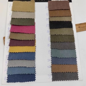 Op de afbeelding: Een kleurenstalenkaart met stofstalen in tinten beige, groen, roze, geel en blauw. De stalen zijn gerangschikt in rijen en kolommen, waarbij elke staal is genummerd. Een houten liniaal is zichtbaar op de achtergrond.