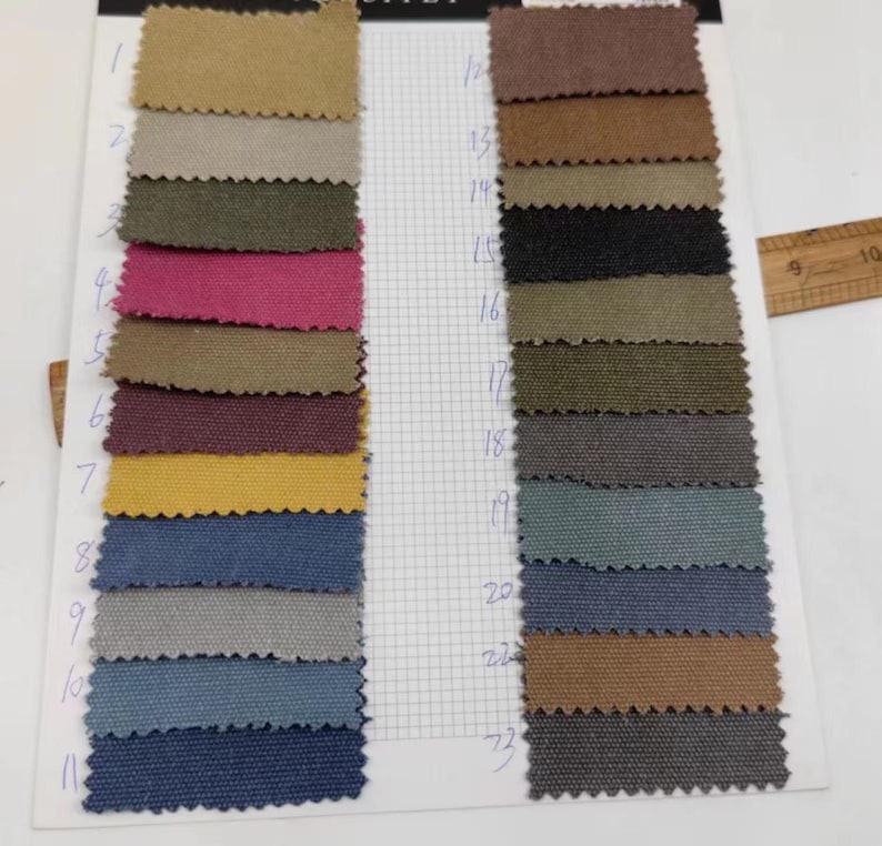 Op de afbeelding: Een kleurenstalenkaart met stofstalen in tinten beige, groen, roze, geel en blauw. De stalen zijn gerangschikt in rijen en kolommen, waarbij elke staal is genummerd. Een houten liniaal is zichtbaar op de achtergrond.