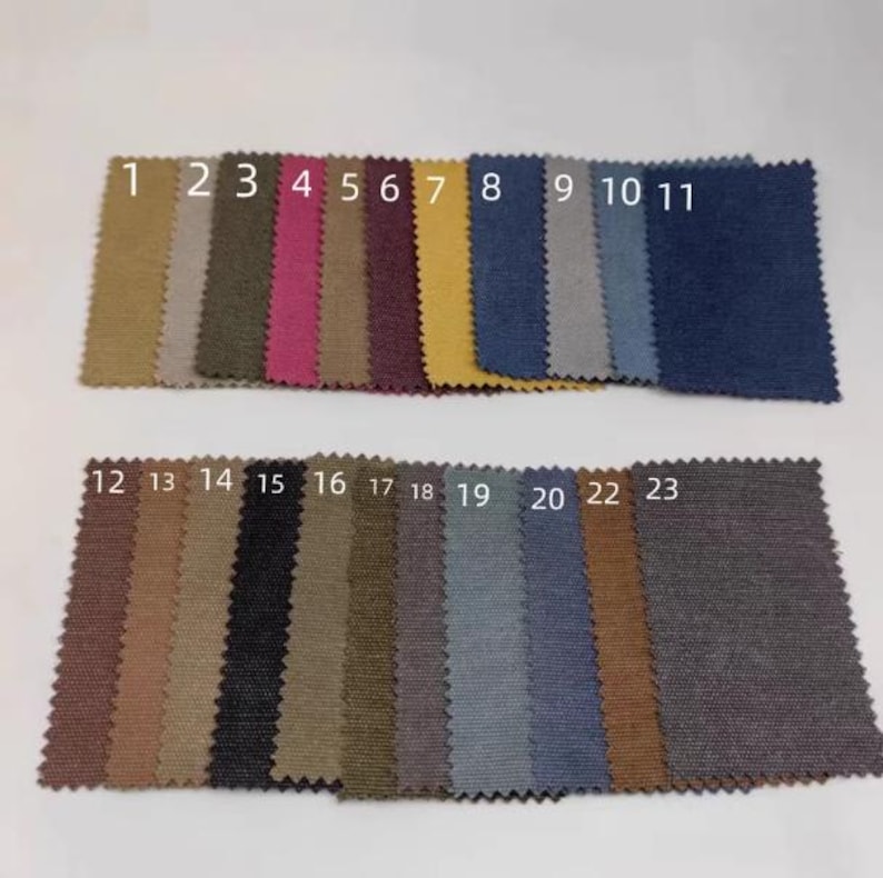 Op de afbeelding: Een verzameling van 23 stoffen stalen in verschillende kleuren, gerangschikt in twee rijen. De kleuren vari&euml;ren van mosterdgeel en dieprood tot marineblauw en antracietgrijs. Elk staal is genummerd van 1 tot 23.