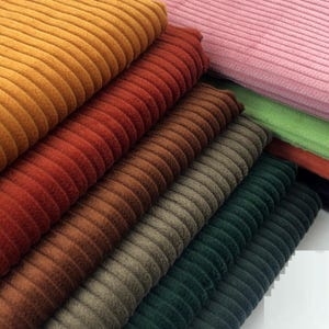 Op de afbeelding: Een verzameling corduroy stofstalen in verschillende kleuren, waaronder oranje, rood, bruin, groen en roze. De stof heeft een geribbelde textuur, waarbij elke kleur netjes op elkaar is gestapeld. De corduroy ziet er zacht en pluche uit.