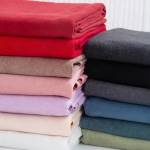 Puede incluir: Montones de tela doblada en varios colores, incluyendo rojo, rosa, gris y azul. Las telas parecen suaves, posiblemente de forro polar o franela. La imagen muestra una variedad de colores y texturas.