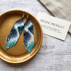 May include: A pair of long, tapered dangle earrings crafted from tiny seed beads in an ombre pattern of black, grey, white, and teal. The earrings feature silver-toned ear wires and are displayed inside a round, crackled gold-toned dish.