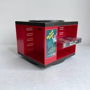 Może przedstawiać: Czerwono-czarny obrotowy pojemnik do przechowywania kaset magnetofonowych. Urządzenie ma prostokątny kształt z wieloma szczelinami na kasety. Kaseta jest widoczna w jednym ze slotów. Na urządzeniu widnieje napis "Die Funk Füchse".