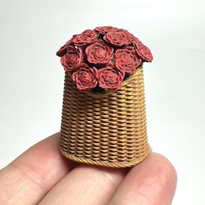 May include: A miniature woven basket filled with red roses. The basket is a light brown color and has a textured, woven pattern. The roses are a deep red and arranged in a dense cluster, creating a charming floral display.