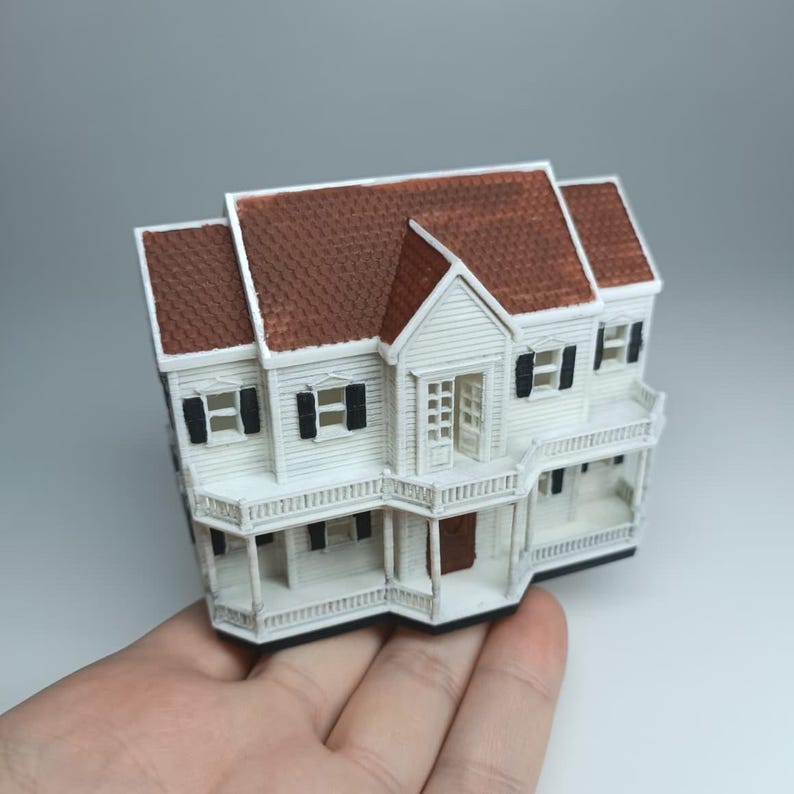 May include: A miniature white house with a brown roof, black shutters, and a porch. The model has detailed architectural features, including a front door and windows. The house is held in a hand against a neutral background.