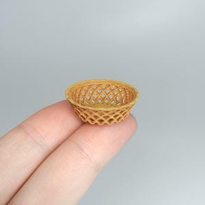Puede incluir: Una pequeña cesta tejida de color marrón claro. La cesta tiene un diseño de celosía y se sostiene entre dos dedos. Es probable que sea un artículo en miniatura o decorativo.