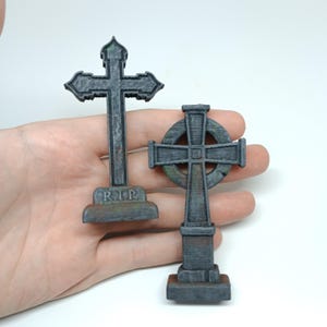May include: Two miniature gray stone crosses with a weathered look. One cross has a circular design and the other has the inscription "R.I.P."