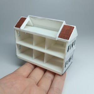 May include: A miniature white two-story house with a brown tiled roof. The house has multiple rectangular compartments and small rectangular window cutouts. The model is held in a hand.