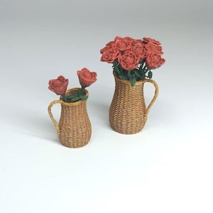May include: Two miniature woven wicker pitchers, one large and one small, each holding red roses. The larger pitcher is filled with multiple roses, while the smaller one has two roses. The pitchers have curved handles and are set against a white background.