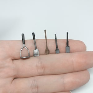 Könnte beinhalten: Ein Set mit sechs Miniatur-Küchenutensilien, darunter ein Kartoffelstampfer, ein Spatel, ein Holzlöffel und drei weitere Werkzeuge. Die Utensilien sind aus Metall und Holz und in Schwarz und Silber lackiert.