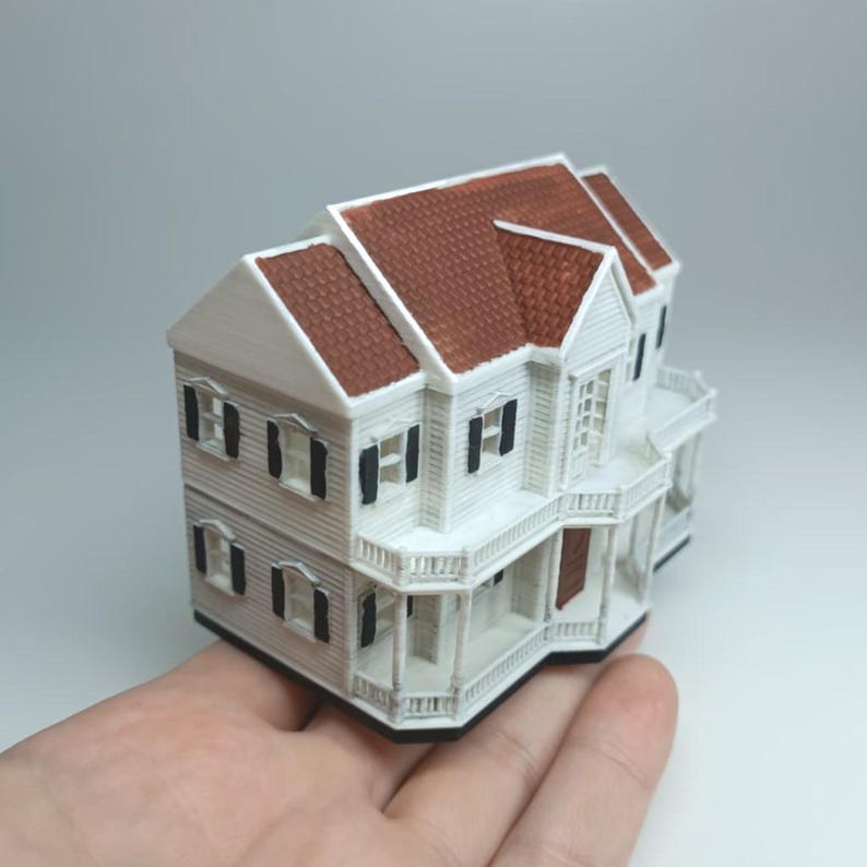 May include: A miniature white house with a brown roof and black shutters. The model has a porch and balcony with intricate railing details. The house is a detailed replica of a classic architectural style.